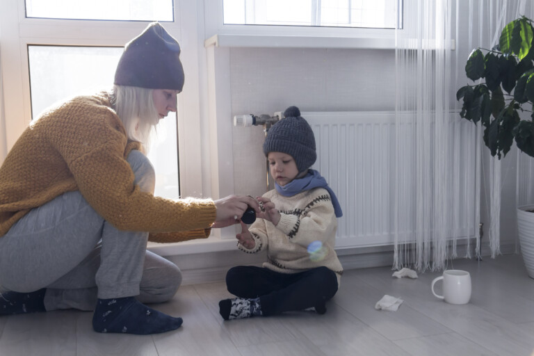 A young mother gives medicine to her little son, who is warmly dressed and sitting on the floor..